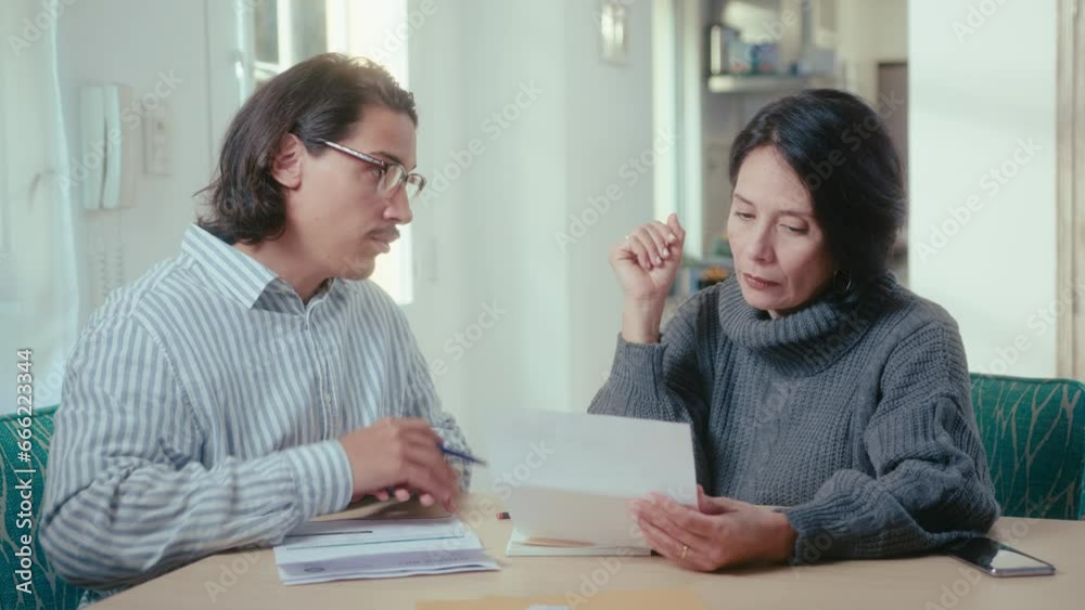 Financial advisor giving documents and explaining information to mature Hispanic woman sitting at table in the living room during consultation at home Financial advisor giving documents and explaining information to mature Hispanic woman sitting at table in the living room during consultation at home