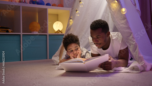 African American man reads fairy tale to preschooler son at night in cozy tent