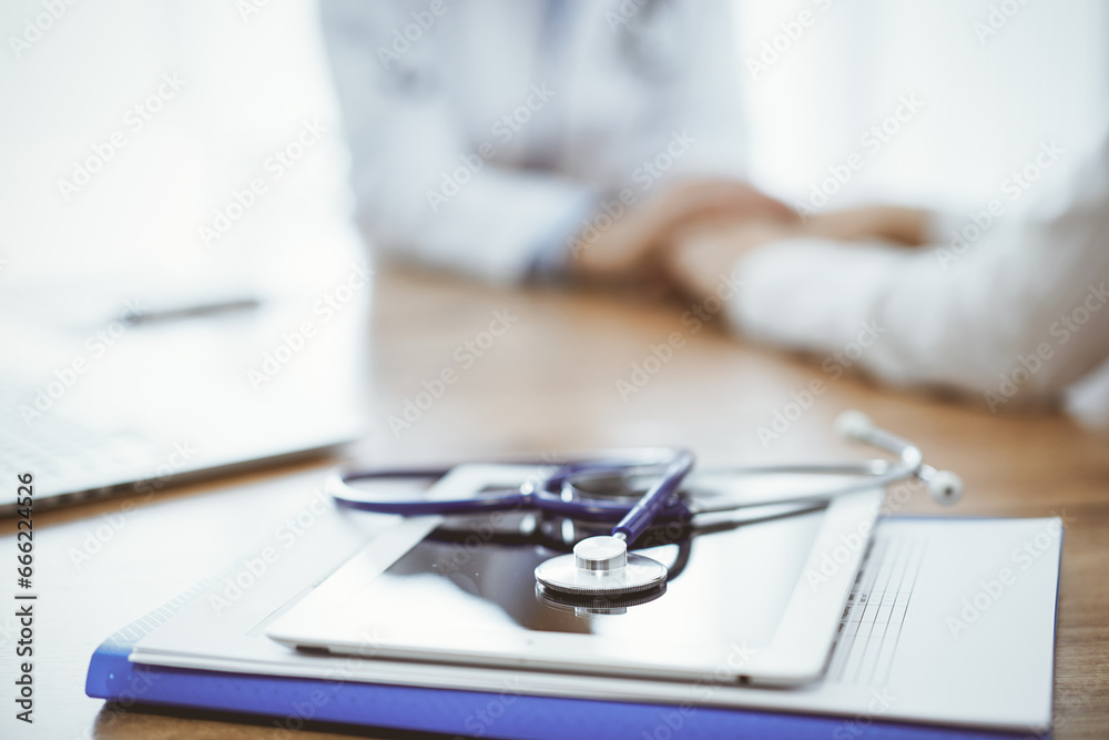 Fototapeta premium Stethoscope, tablet and laptop computer are lying on the wooden table while doctor's hands reassuring a patient at the background. Medicine concept
