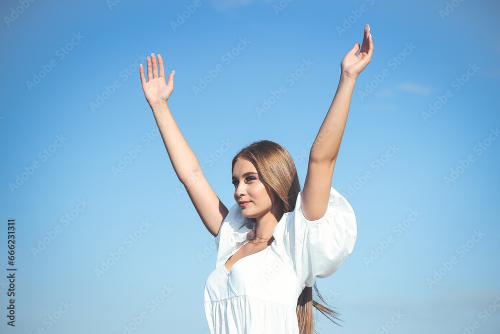 Happy, beautiful woman on the ocean beach standing in a white summer dress, raising hands