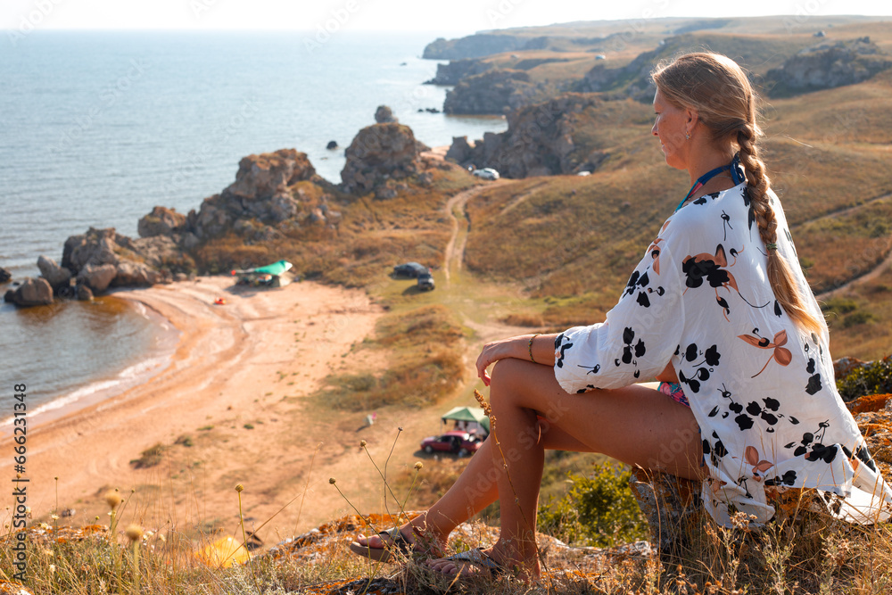 A mature woman with a braid sits on a stone on the top of a mountain and looks at the sea coast with a tourist camp. Travel and tourism