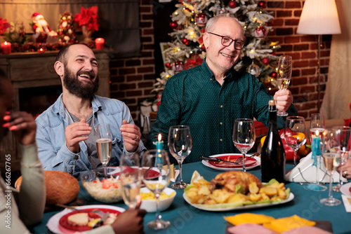 Senior man doing toast with raised glass of wine at christmas eve dinner to celebrate december holiday with friends and family. Grandfather making speech around the table  sparkling alcohol.