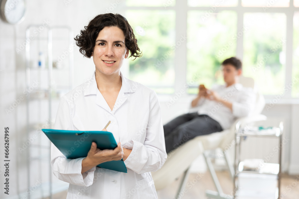 Glad middle-aged woman doctor standing with folder against background of patient lying on clinical chair