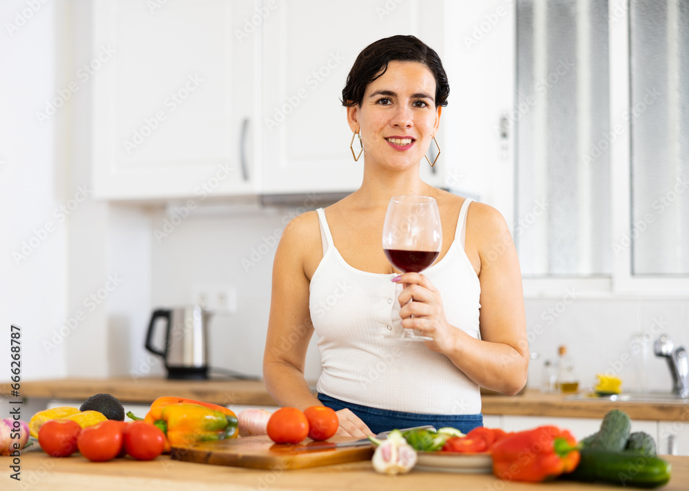 Portrait of pleased woman with glass of red wine in modern kitchen - housewife prepares a vegetable salad and drinks red wine