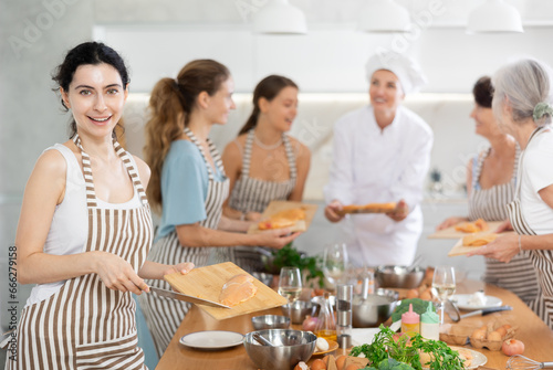Young woman holding cutting board with raw chicken breast in her hands posing surrounded by other members of cooking course