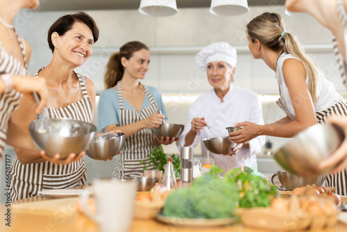 Aged woman holding bowl and whisk in her hands and talking to other female members during cooking master class photo