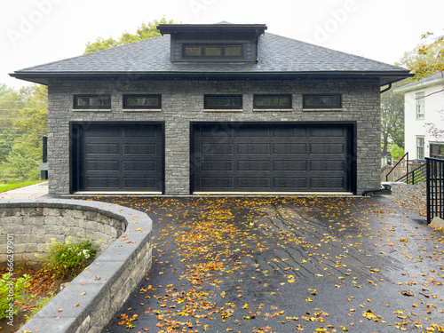 A double and single-width black metal garage door with panels in a grey brick luxurious residential outcrop building. There are small glass windows above the doors with a dormer window on the roof. 