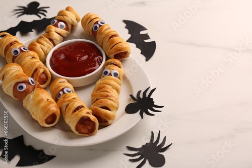 Plate with tasty sausage mummies, Halloween decorations and ketchup on white table, closeup. Space for text