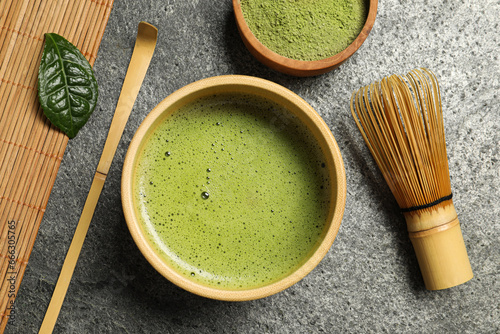 Cup of fresh matcha tea, bamboo whisk, spoon and green powder on grey table, flat lay
