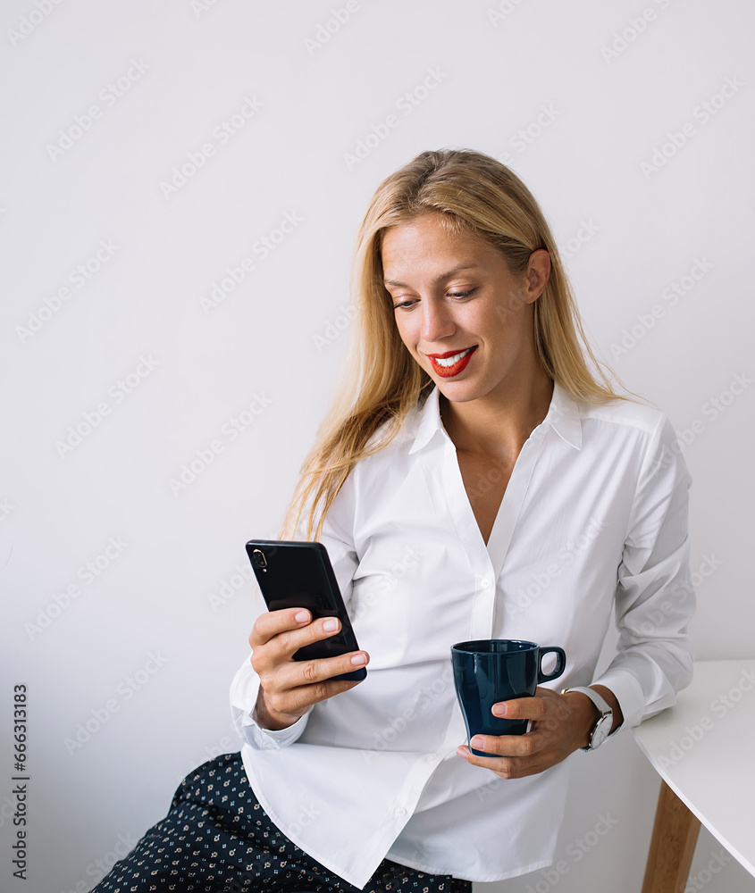 Serious female sitting at table with blue mug messaging on phone and working on laptop Serious female sitting at table with blue mug messaging on phone and working on laptop