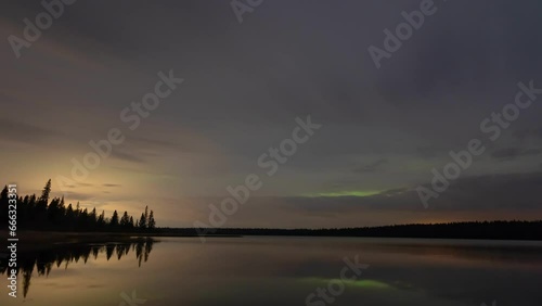 Gray nighttime clouds with the occasional colorful patch of Aurora move towards the viewer above a calm lake in theis time lapse. The scene is near perfectly reflected in the water.
 photo