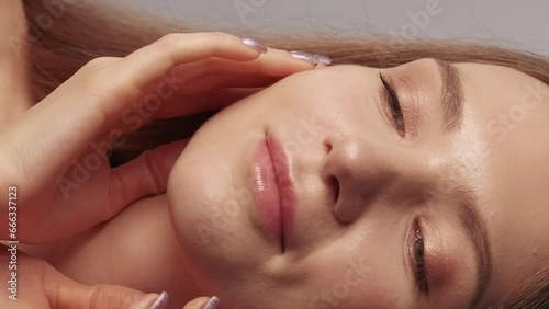 Vertical video. Face moisturizing. Skincare dermatology. Pretty young woman with nude makeup enjoying touching soft flawless perfect radiant shiny skin. photo