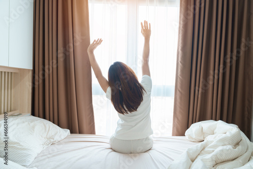 happy woman stretching on bed after wake up, young adult female rising arms and looking to window in the early morning. fresh relax and have a nice day concepts