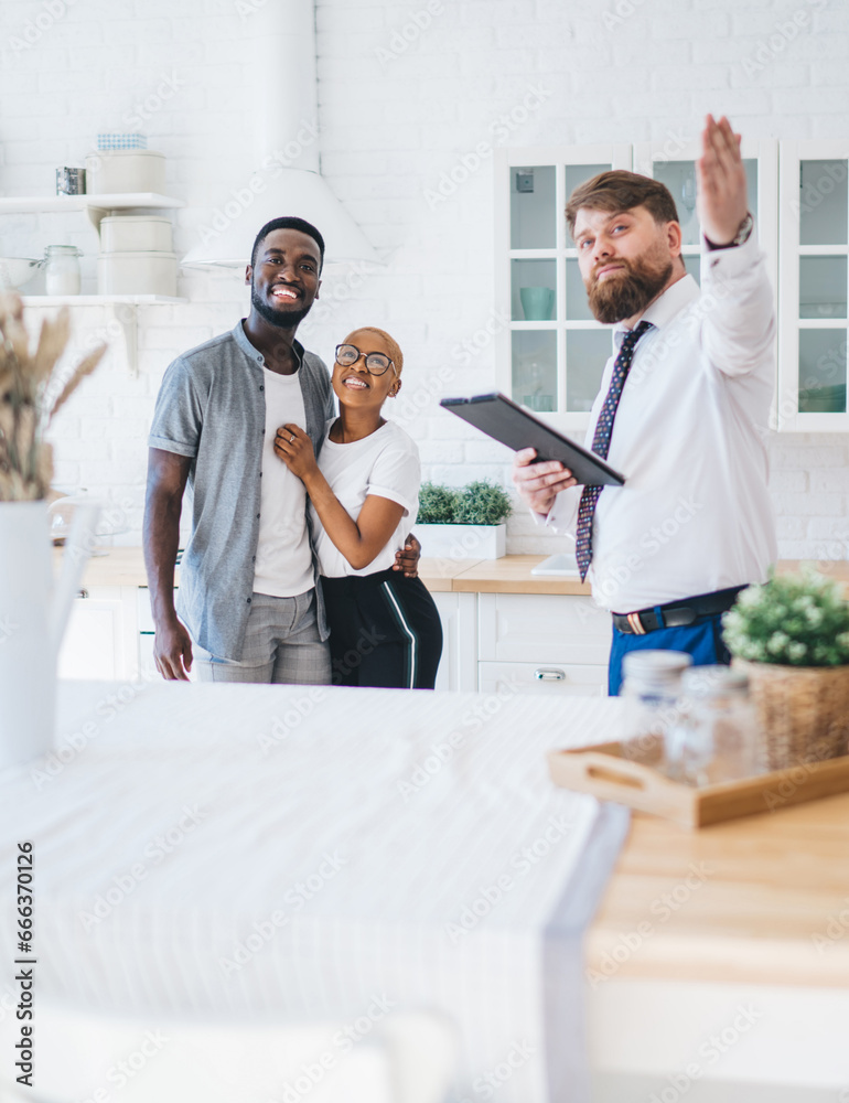 © BullRun - Confident estate agent showing kitchen to African American pleased hugging couple