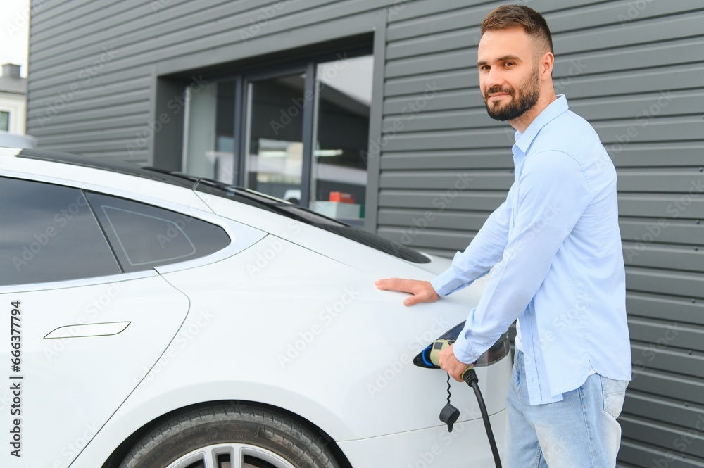 Beautiful young stylish man is with electric car at daytime charging the vehicle