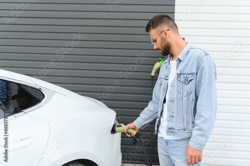 Man Holding Power Charging Cable For Electric Car In Outdoor Car Park. And he s going to connect the car to the charging station in the parking lot near the shopping center. © Serhii