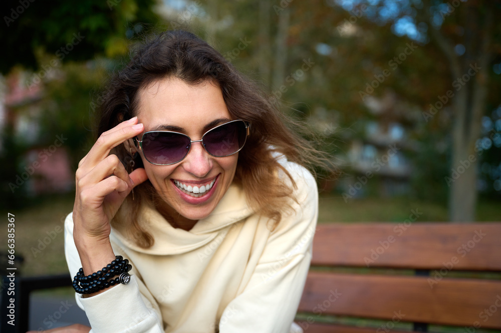Portrait of a young woman in a modern sun glasses sitting on the wooden bench in a park near a lake enjoying the sunset.Stylish girl in light sports clothes sitting and relaxing on the beach at sunset Portrait of a young woman in a modern sun glasses sitting on the wooden bench in a park near a lake enjoying the sunset.Stylish girl in light sports clothes sitting and relaxing on the beach at sunset