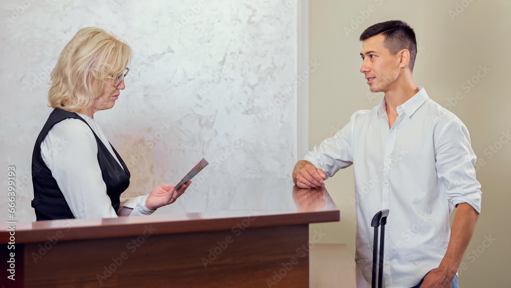 Obraz premium Young man handing over passport to find reserved room in hotel. Blonde female receptionist checking visitor reservation in computer