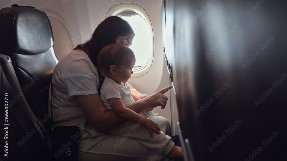 Mother caresses and playfully engages with toddler girl. Mom holds child securely in arms as family soars through skies on journey by plane Mother caresses and playfully engages with toddler girl. Mom holds child securely in arms as family soars through skies on journey by plane