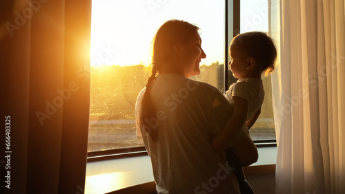 Mom holds toddler girl in arms approaching window. Mother tenderly shows vibrant sunset to curious child sharing moment of warmth and wonder © lenblr