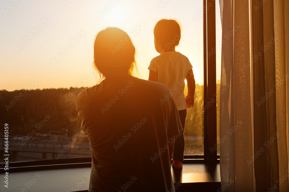 Perched on windowsill toddler girl under supervision of mother looks through window at picturesque sunset. Colors of sun give tenderness to picture, sunlight