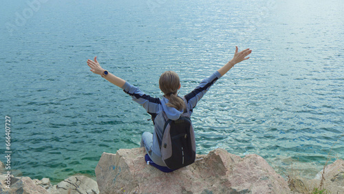 Female tourist with backpack on her shoulders sits on rock above sea with her hands raised up.