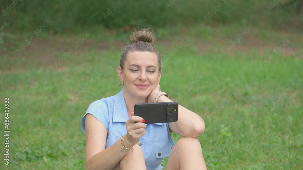 Positive woman looks at screen of her smartphone while sitting on grass in park. She enjoys and smiles. Positive woman looks at screen of her smartphone while sitting on grass in park. She enjoys and smiles.