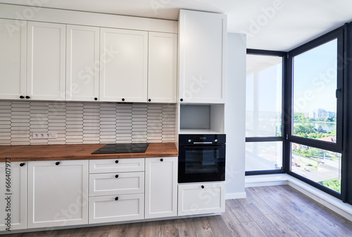 Kitchen with laminate floor, white walls, doorway and modern kitchen set with electric stove, counter, sink and oven. Apartment kitchen room with elegant minimalist design in white colors.