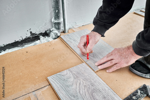 Close up of man construction worker with pencil in hand drawing mark on laminate wooden board. Male worker preparing laminate material for floor installation in apartment under renovation.