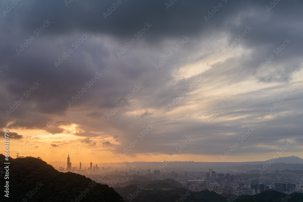 At dusk, the sun breaks through the clouds and shines on Taipei City. Silhouettes of city buildings. Orange sky and dark clouds.