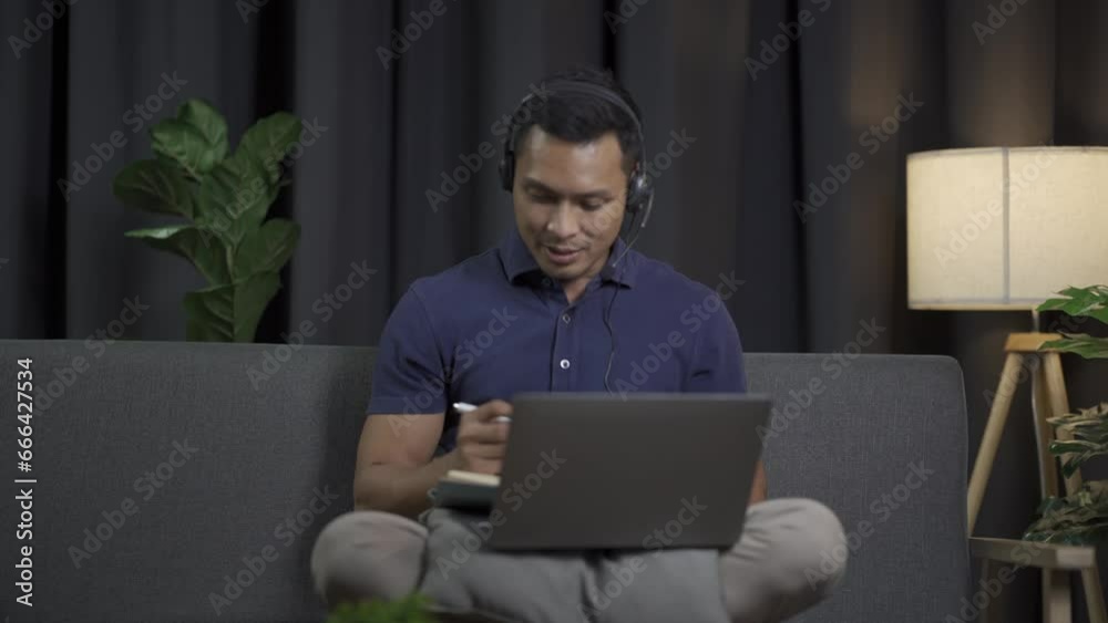 A young black-haired man sits on a sofa and works using his laptop. Young man working remotely Man wearing headphones watching movies, listening to music at home on a casual day. A young black-haired man sits on a sofa and works using his laptop. Young man working remotely Man wearing headphones watching movies, listening to music at home on a casual day.