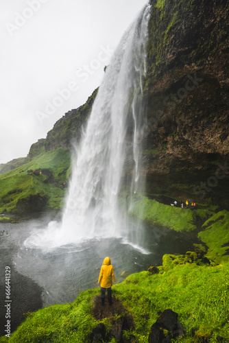 View of a girl in a yellow rain coat standing on a rock below the Seljalandsfoss waterfall in Southern region of Iceland.