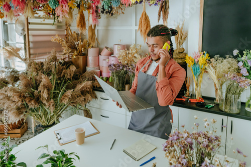 Young entrepreneur talking on smart phone and using laptop in flower shop photo