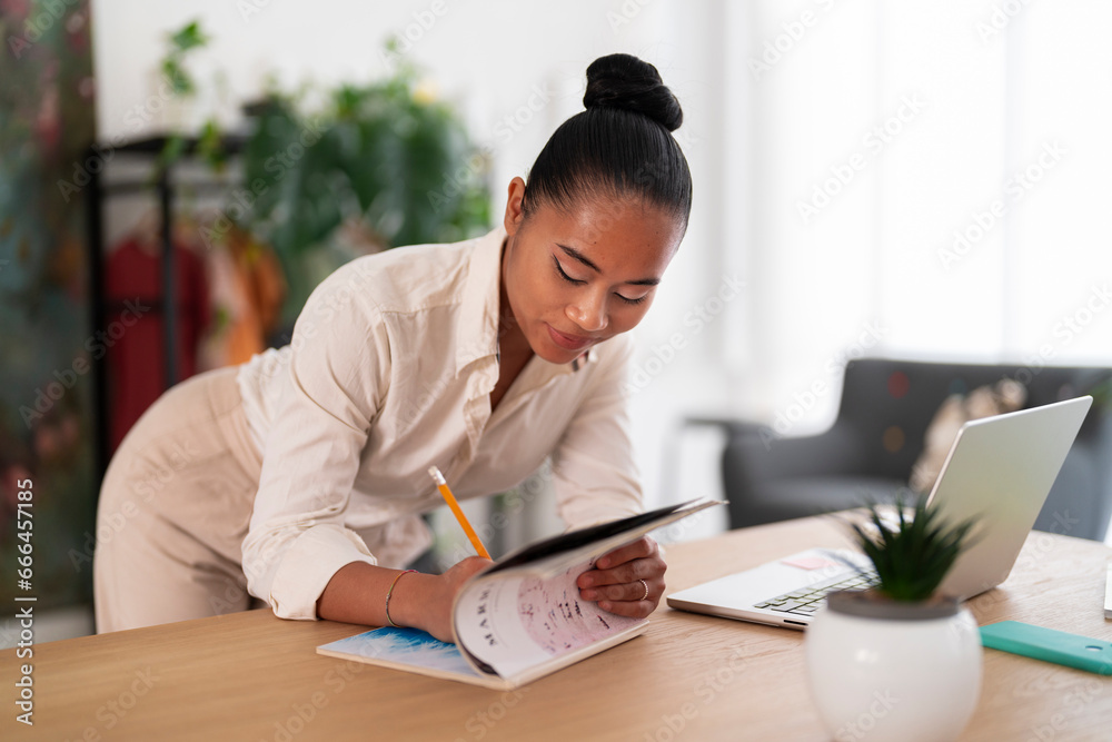 Young Asian female employee in smart casual outfit with hair bun leaning forward while writing notes in magazine during workday in modern office Young Asian female employee in smart casual outfit with hair bun leaning forward while writing notes in magazine during workday in modern office