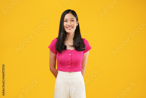 A smiling young Asian woman with straight hair is posing to the camera standing and isolated on a yellow background in a studio
