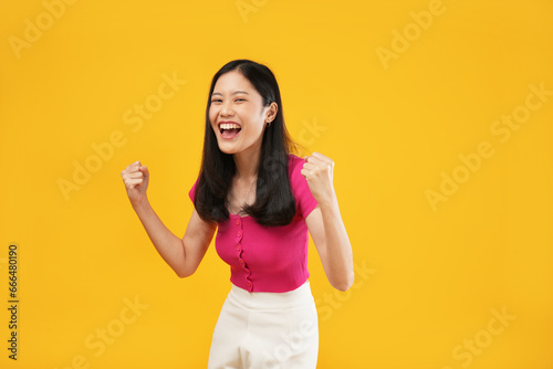 Photo of a cheerful young Asian woman her hands clenched into fists and a happy smile, isolated on a yellow background. 