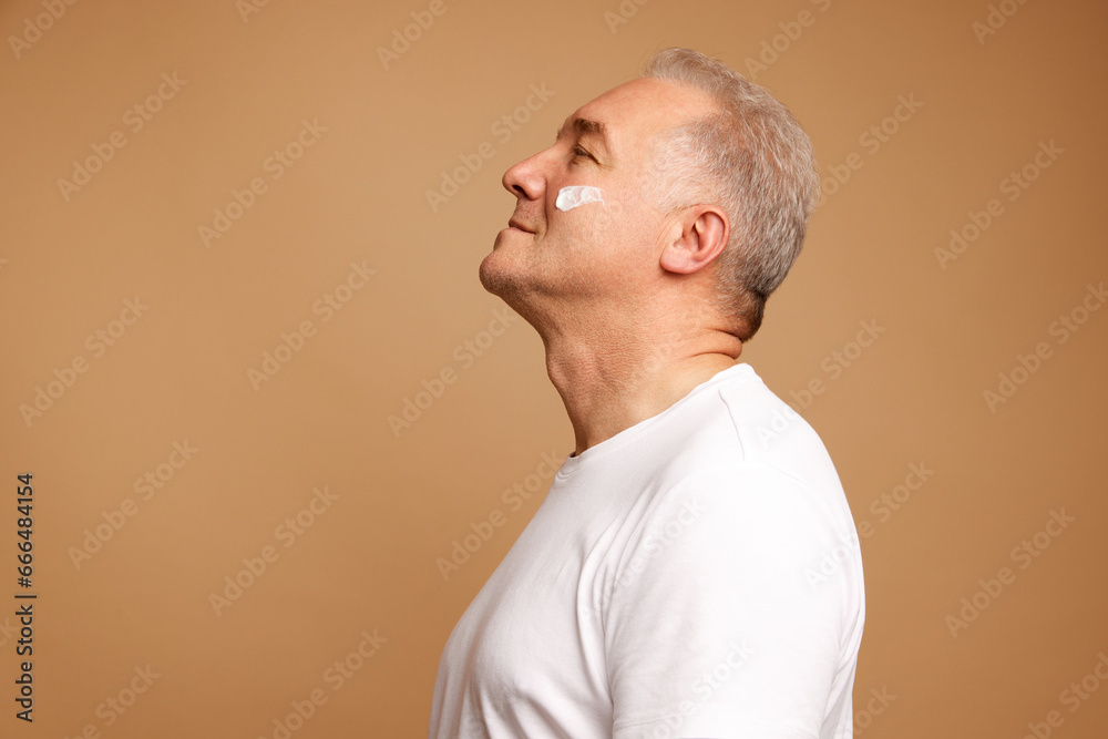 Side view portrait of middle aged, 45s man with cream, lotion swatches on face against pastel beige background. Side view portrait of middle aged, 45s man with cream, lotion swatches on face against pastel beige background.