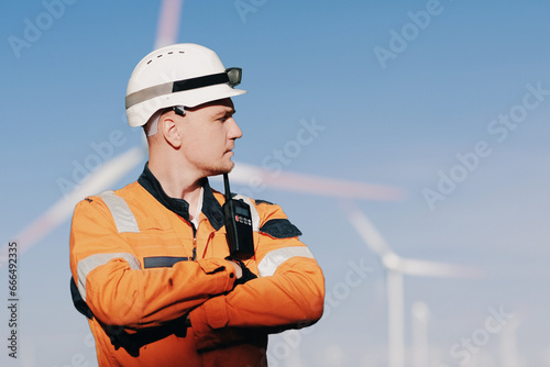 Wind Farm Offshore Maintenance Technician. Seafarer. Seaman. Navigator. A Man In A Working Overall Boiler Suit With A Radio And Safety Helmet With A Blurred Wing Generators In The Background