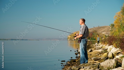 Full length view of a mature adult man on the river bank, fishing with a spinning rod. A man in jeans and a shirt enthusiastically spins the reel on his fishing rod, dreaming of catching a big fish.
