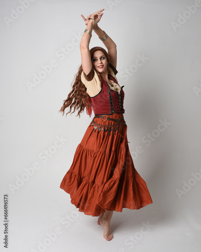 Full length portrait of beautiful red haired woman wearing a medieval maiden, fortune teller costume. Standing pose with dancing gestures, twirling skirt. isolated on studio background.