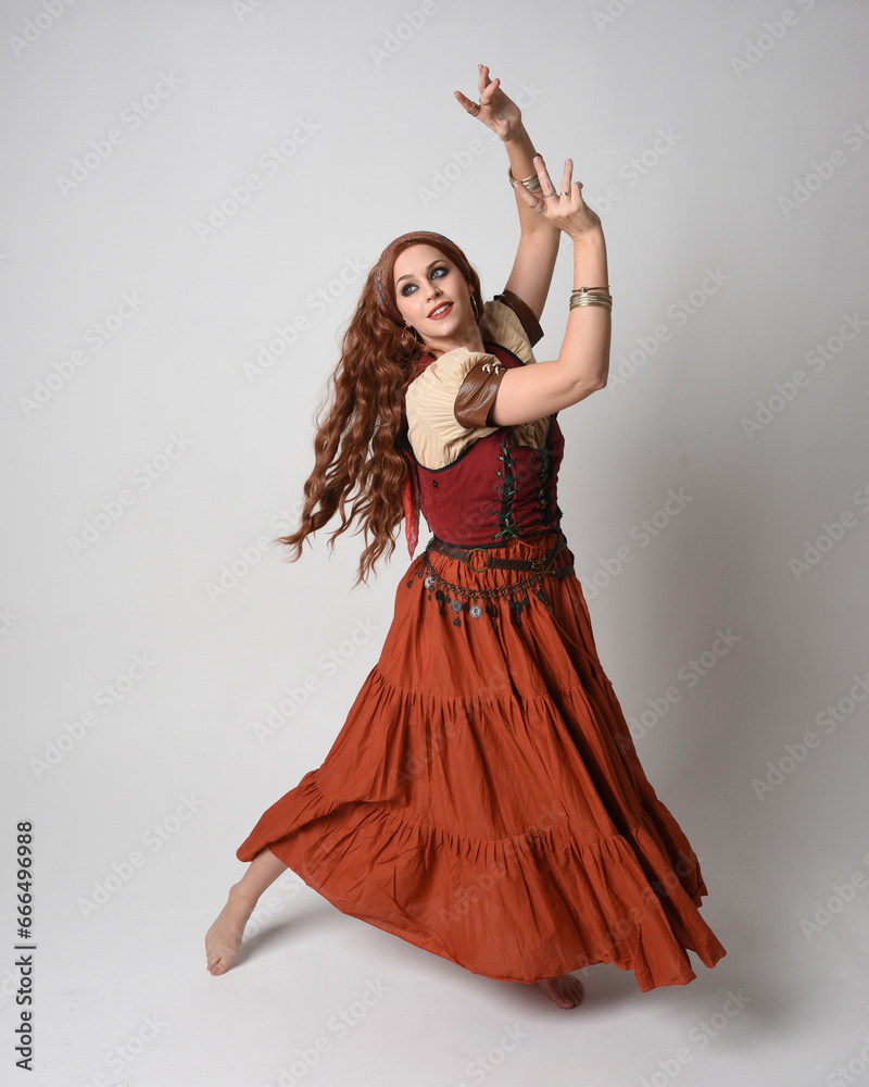 Full length portrait of beautiful red haired woman wearing a medieval maiden, fortune teller costume. Standing pose with dancing gestures, twirling skirt. isolated on studio background.