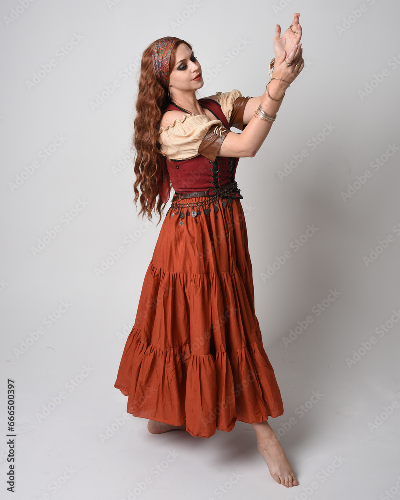 Full length portrait of beautiful red haired woman wearing a medieval maiden, fortune teller costume. Standing pose with dancing gestures, twirling skirt. isolated on studio background.