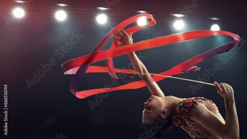Portrait of a Graceful Rhythmic Gymnast Performing a Dance with a Red Ribbon. Young Female Exercising in a Striking Beautiful Uniform in a Dark Studio. Cinematic Super Slow Motion Speed Ramp Footage