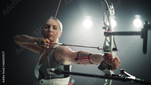 Cinematic Portrait of a Professional Female Archer Aiming and Shooting an Arrow at a Target with a Modern Compound Bow. Strong Athlete is Fully Equipped and Protected with Chest and Arm Guards photo