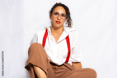 Young brunette woman with curly black hair wearing a white shirt, black lingerie, red suspenders and light brown pants