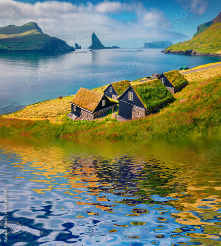 Bour village with typical turf-top houses reflected in the calm waters of small pond. Stunning summer scene of Tindholmur cliffs on Vagar island, Faroe, Denmark, Europe. Traveling concept background.. photo