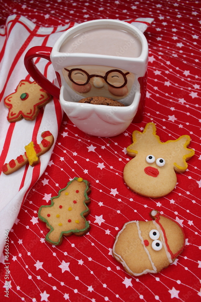 Fototapeta premium A Santa Claus-shaped mug filled with hot chocolate or coffee. A chocolate chip cookie peeks out of the cup. Homemade Christmas cookies. Red tablecloth with white stars and a striped kitchen towel. 