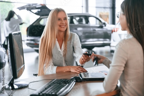 Giving the keys. Female manager is helping woman customer in the car dealership salon