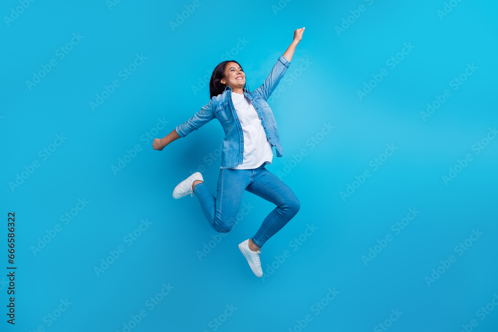 Full length photo of optimistic person wear denim jacket pants flying raising fist look empty space isolated on blue color background