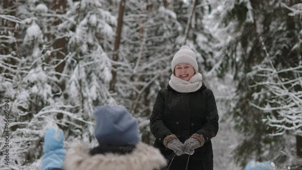 Portrait of happy nanny carries child on sled and cutely waves her hand in mitten to him in snowy forest. Woman and child walk in warm clothes through fabulous winter forest on vacation. Portrait of happy nanny carries child on sled and cutely waves her hand in mitten to him in snowy forest. Woman and child walk in warm clothes through fabulous winter forest on vacation.